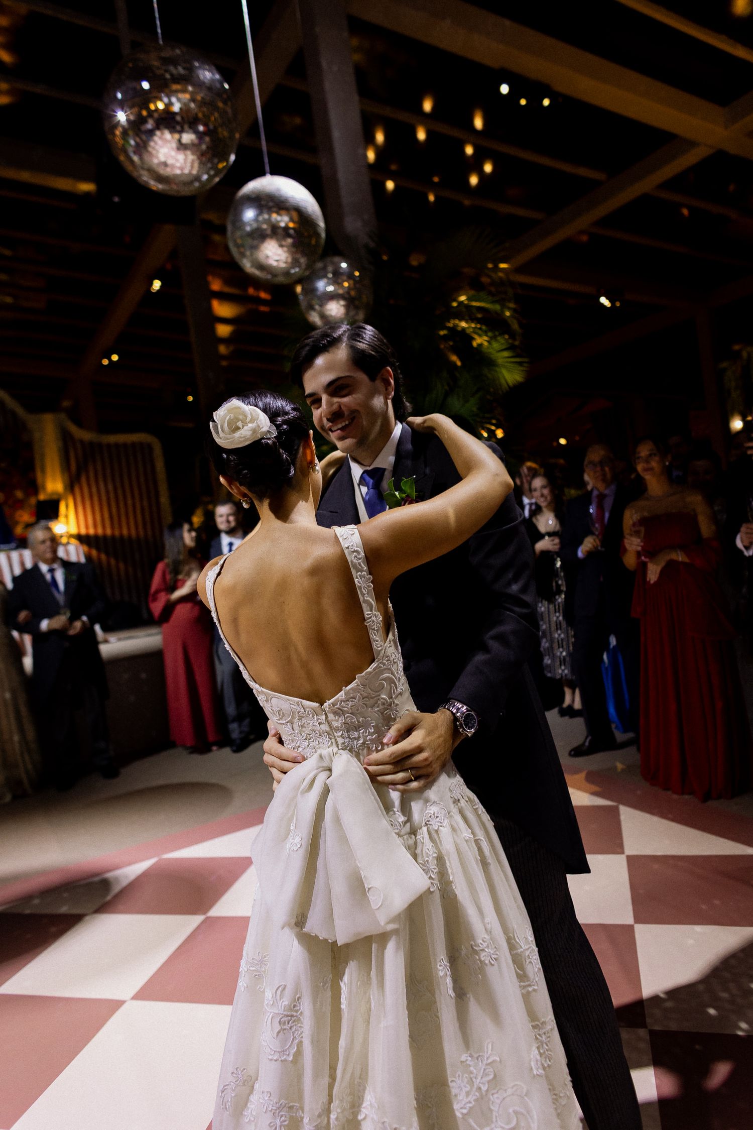 A couple dances together at a wedding reception under disco balls and dim lighting, with the bride wearing a white backless gown.