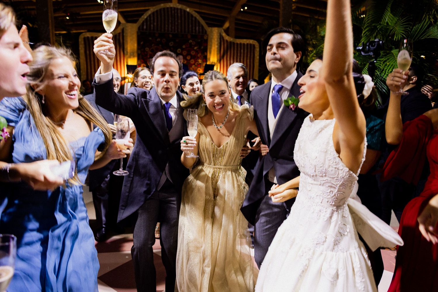 Wedding guests dance and celebrate on a lively dance floor at an evening reception with champagne glasses raised.