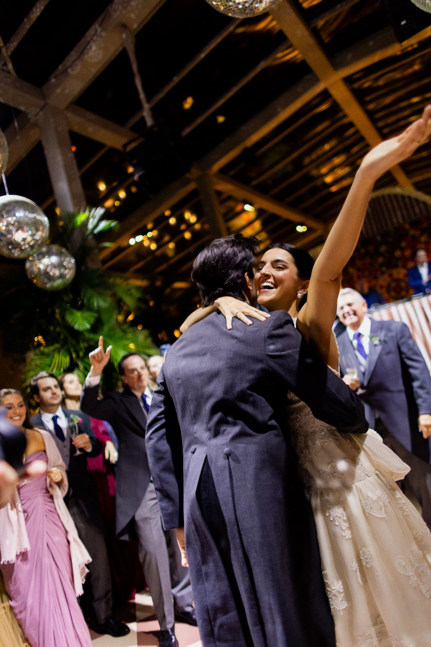 A couple shares their first dance at a wedding reception under disco balls and warm lighting while guests look on.