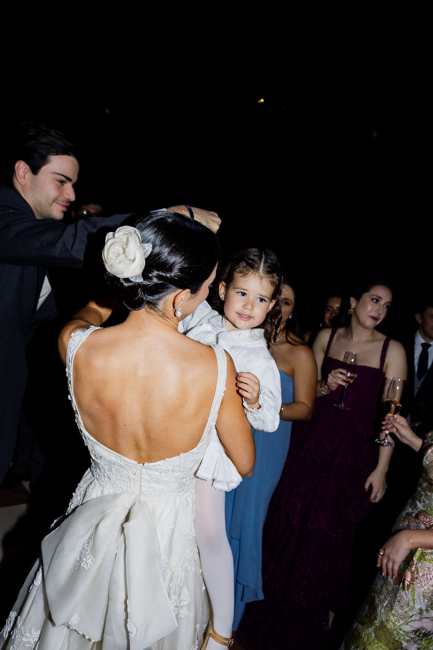 A small child in a white dress being carried at a formal event with guests in purple and blue dresses in the background.
