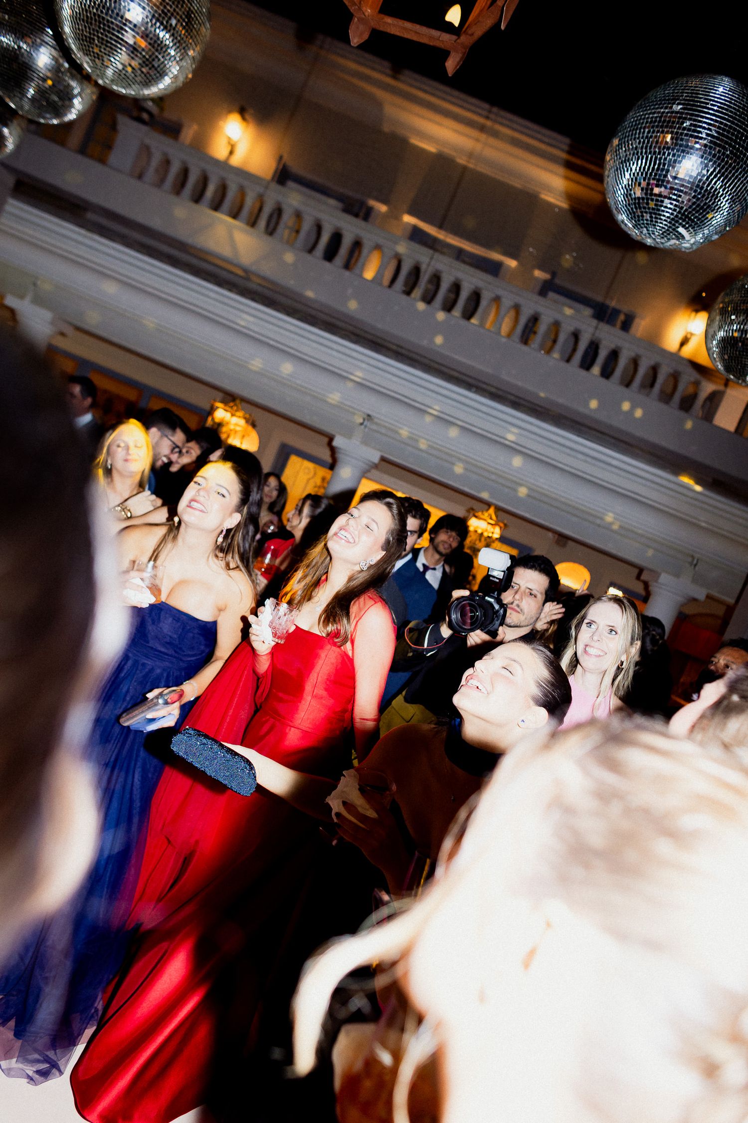 Disco balls hang above a crowded dance floor at an elegant formal event with partygoers in evening attire.
