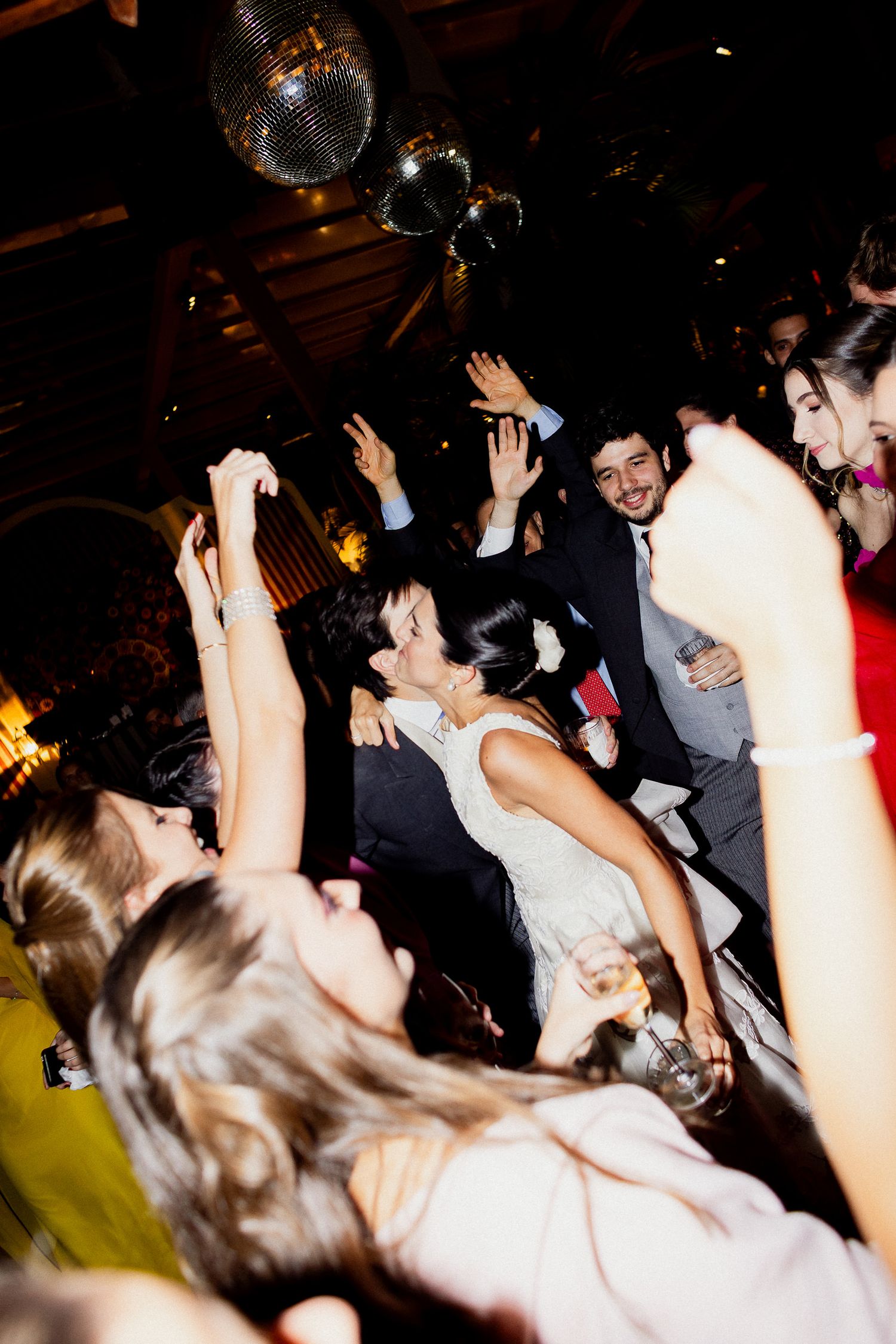 A crowd of wedding guests dancing with their hands raised in celebration at a dimly lit reception venue.
