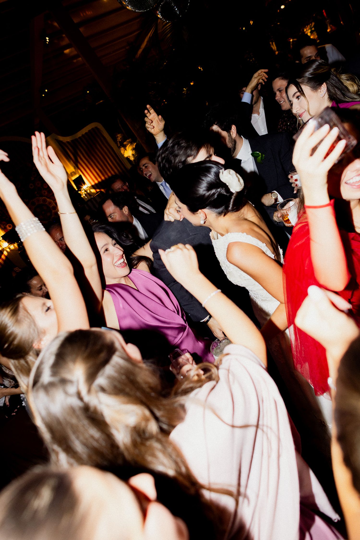Energetic crowd of people dancing with raised arms at a dark nightclub or party venue.