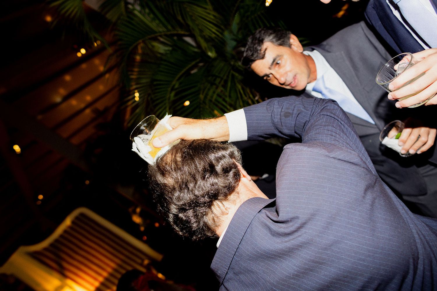 Two people in formal attire dancing at an evening event with dim lighting and palm trees in the background.