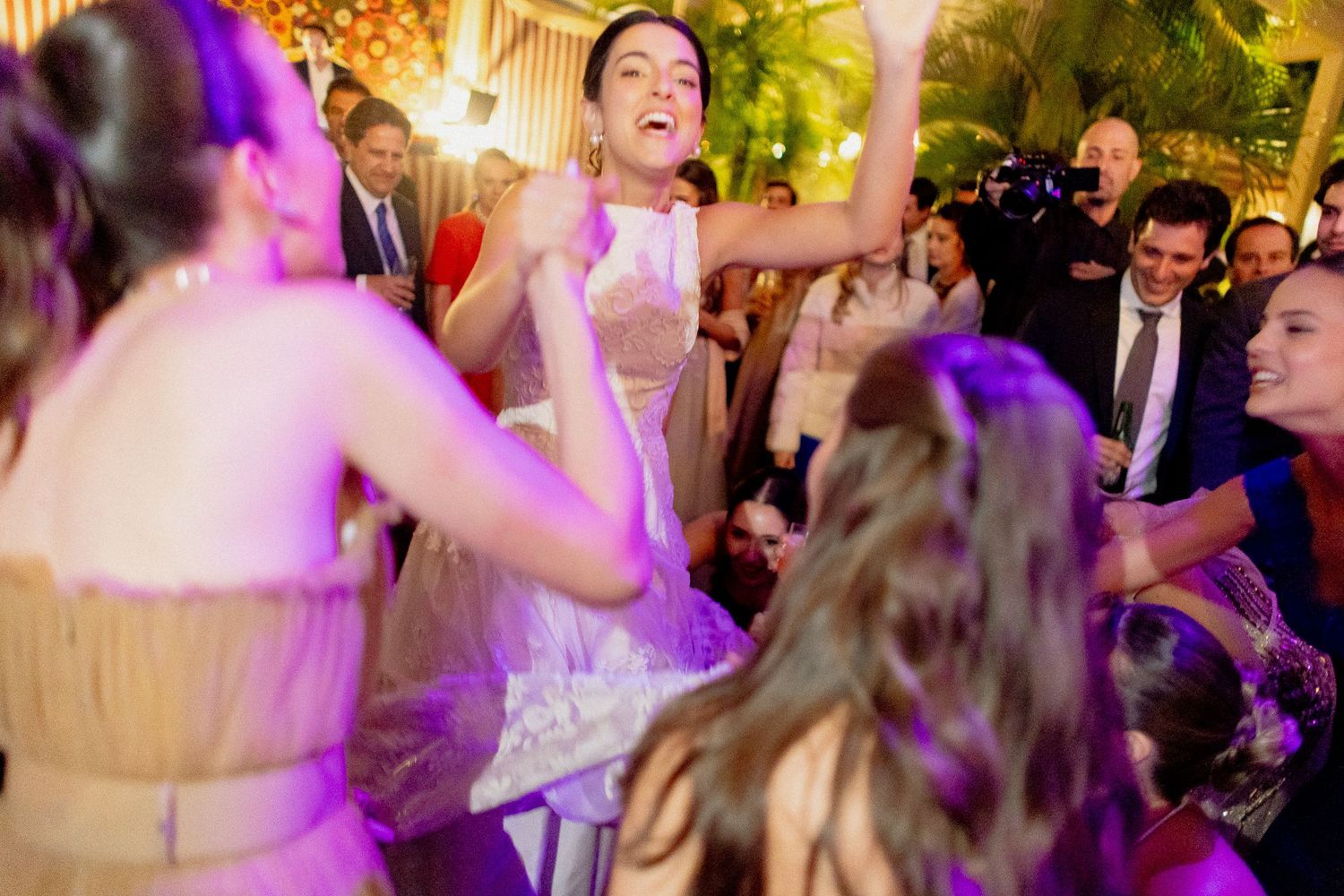 Wedding guests dance and celebrate on a crowded dance floor illuminated by colorful purple and amber lighting.