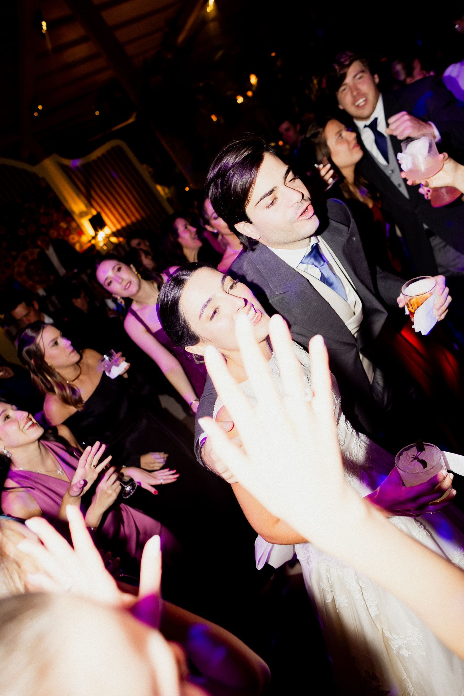 Wedding guests dance energetically on a dimly lit dance floor with purple lighting at an evening reception celebration.