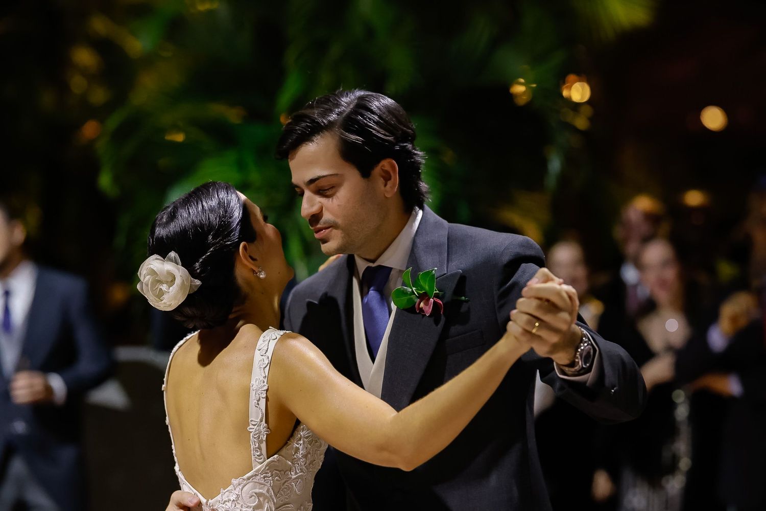 A couple dances together at an elegant evening wedding reception with soft lighting and greenery in the background.