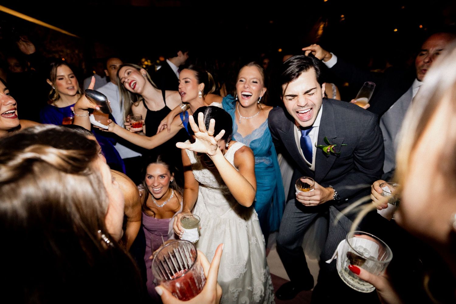 Wedding guests dance enthusiastically on a dark dance floor during a reception celebration.
