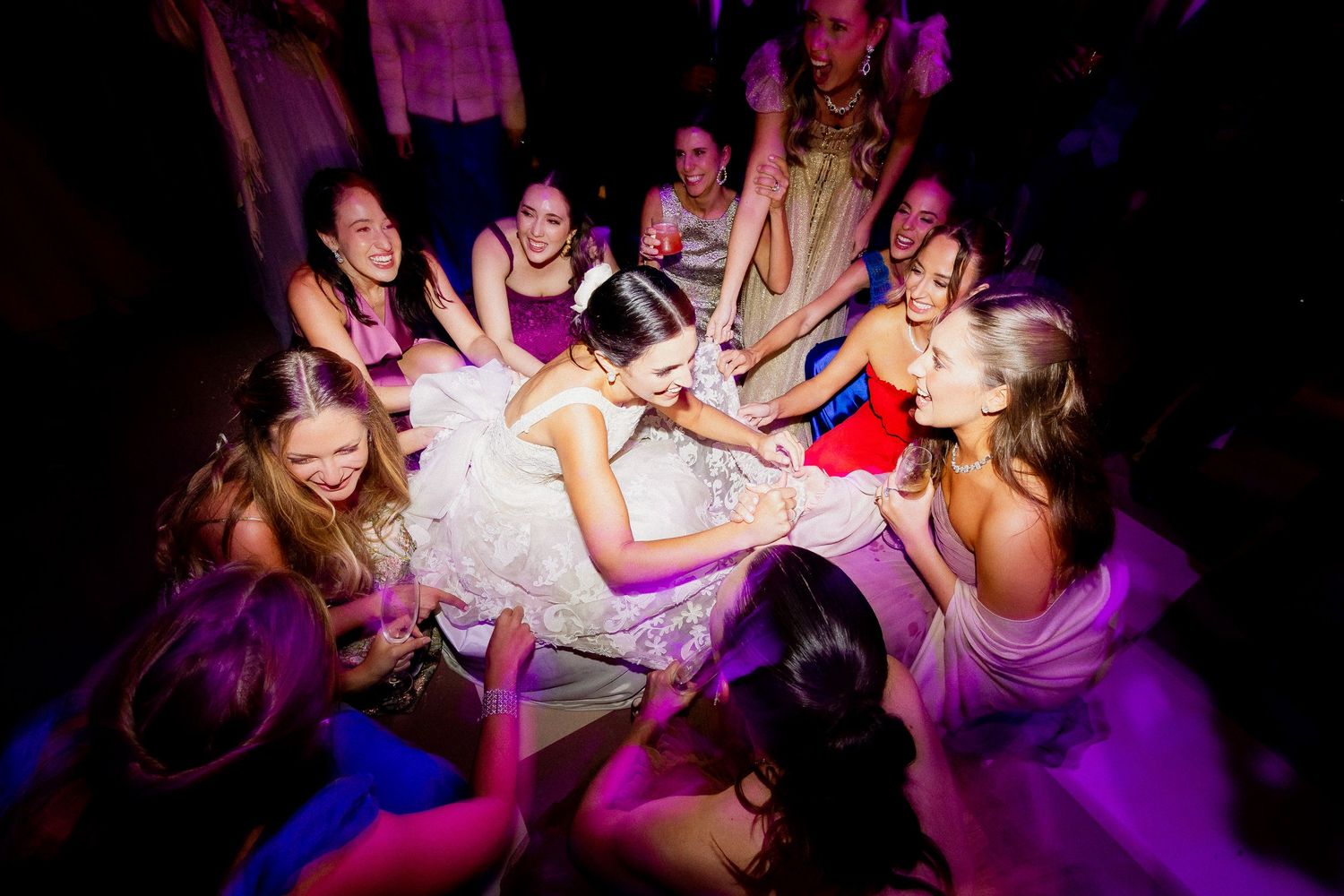 A group in purple dresses gather around a white dress on a dark dance floor during a celebration.
