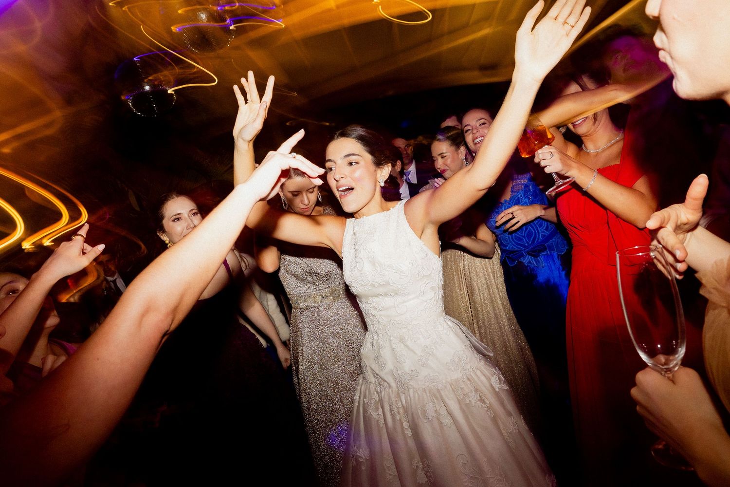Wedding reception guests dance energetically on a crowded dance floor under colorful party lighting.