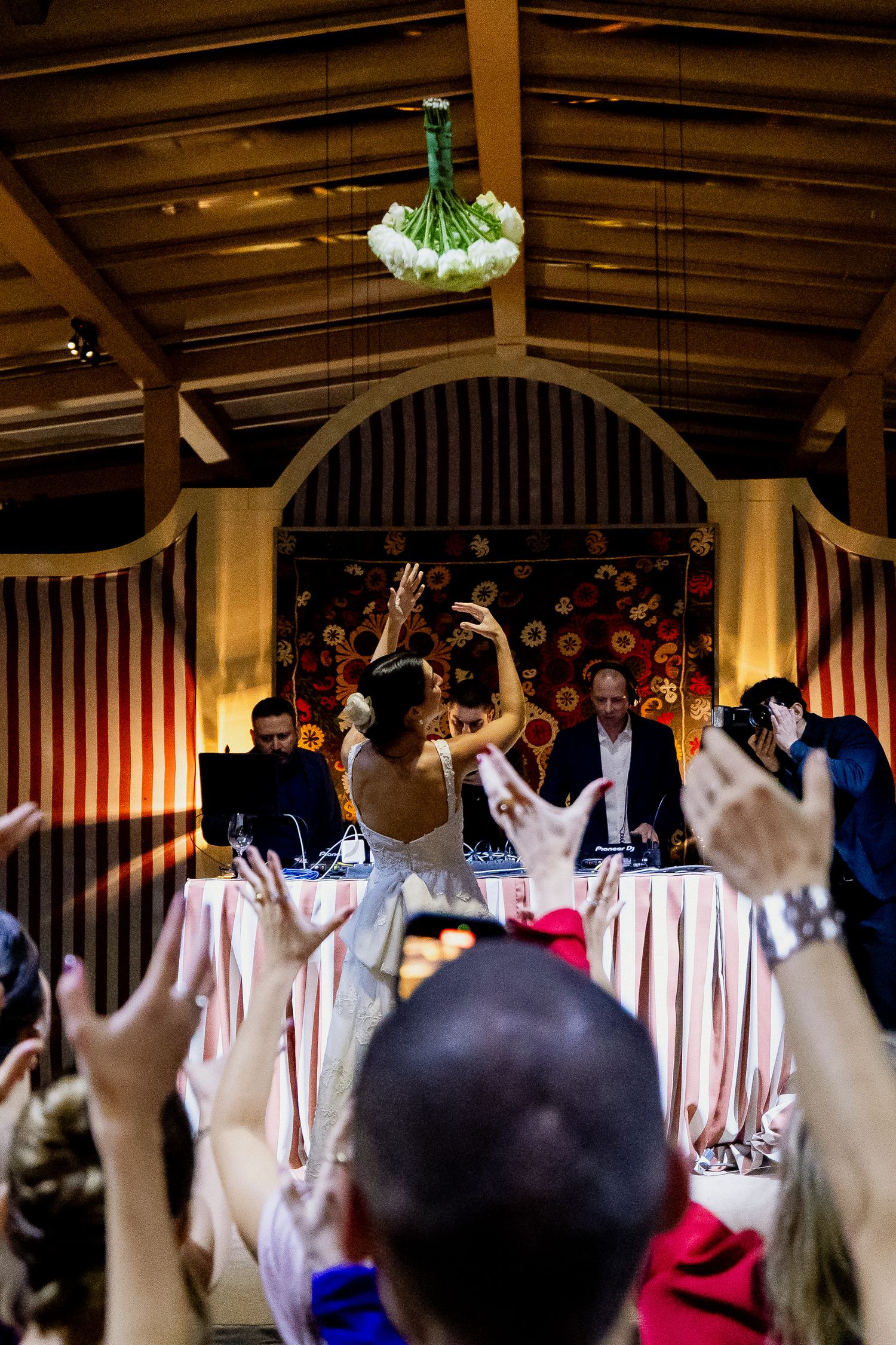 Celebratory moment at a wedding reception with hands raised under wooden beams and hanging floral decorations.