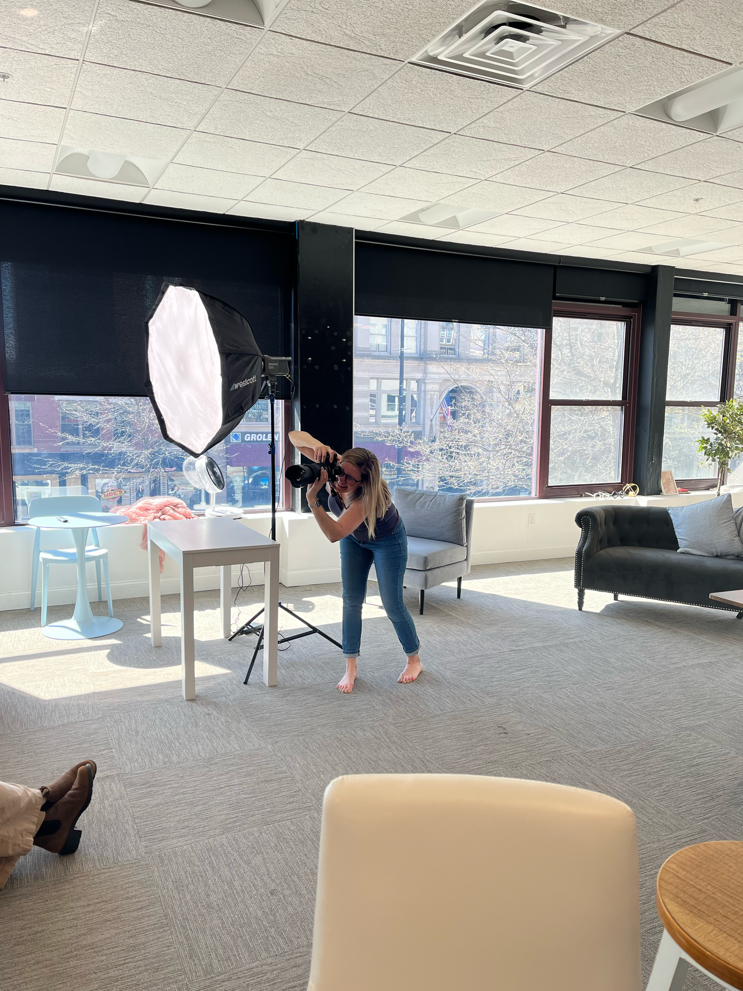 Photographer with camera and softbox light stand shooting in a modern office space with large windows.