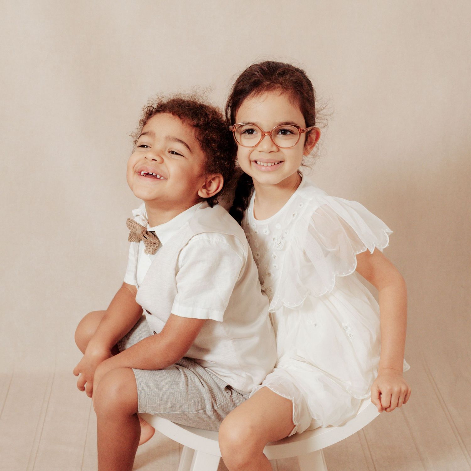 Two school children laughing sitting on a white stool in a studio location with a cream background.
