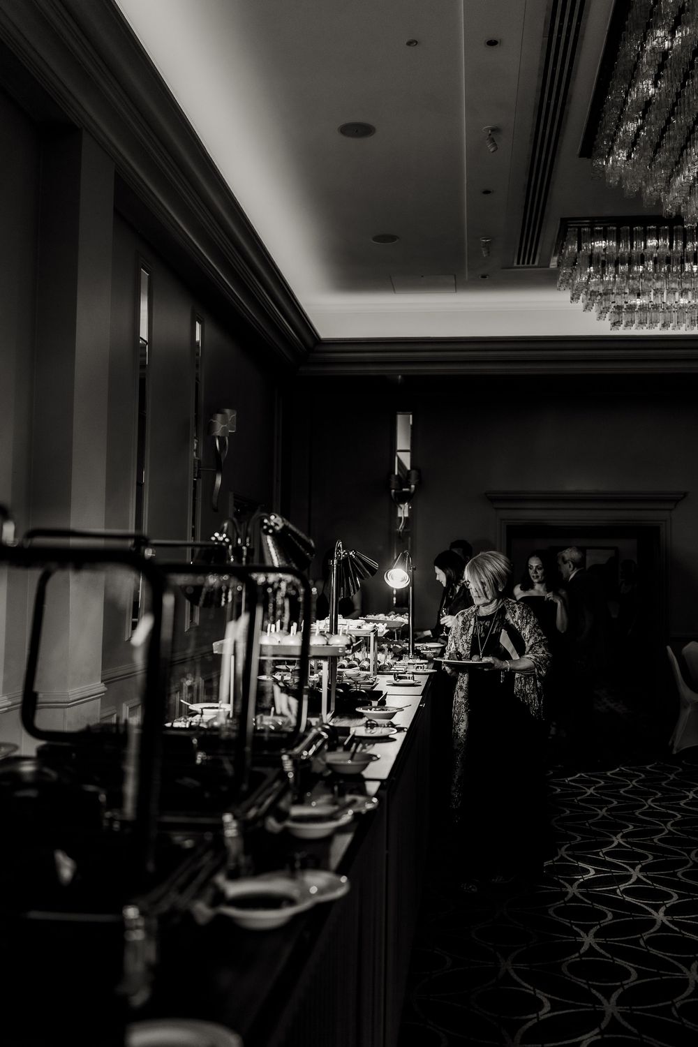 Dark and moody reception hall setup with elegant table settings and crystal chandeliers casting dramatic shadows.