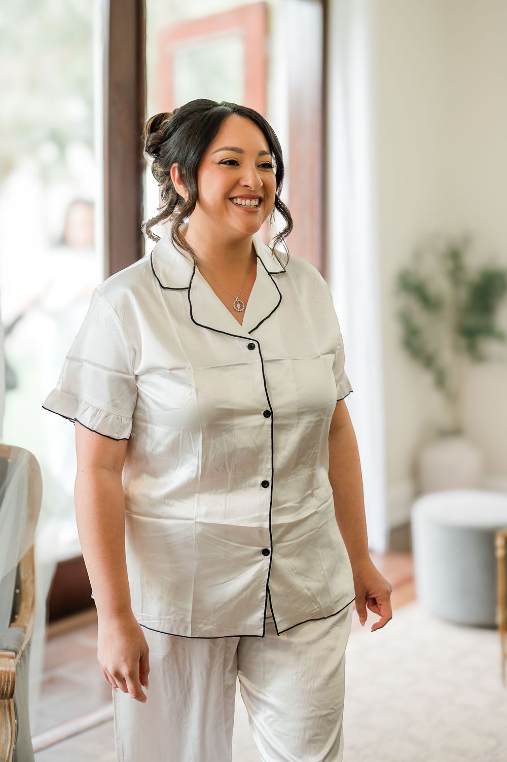 A person wearing white pajamas with black piping trim smiles while sitting in a bright interior setting.