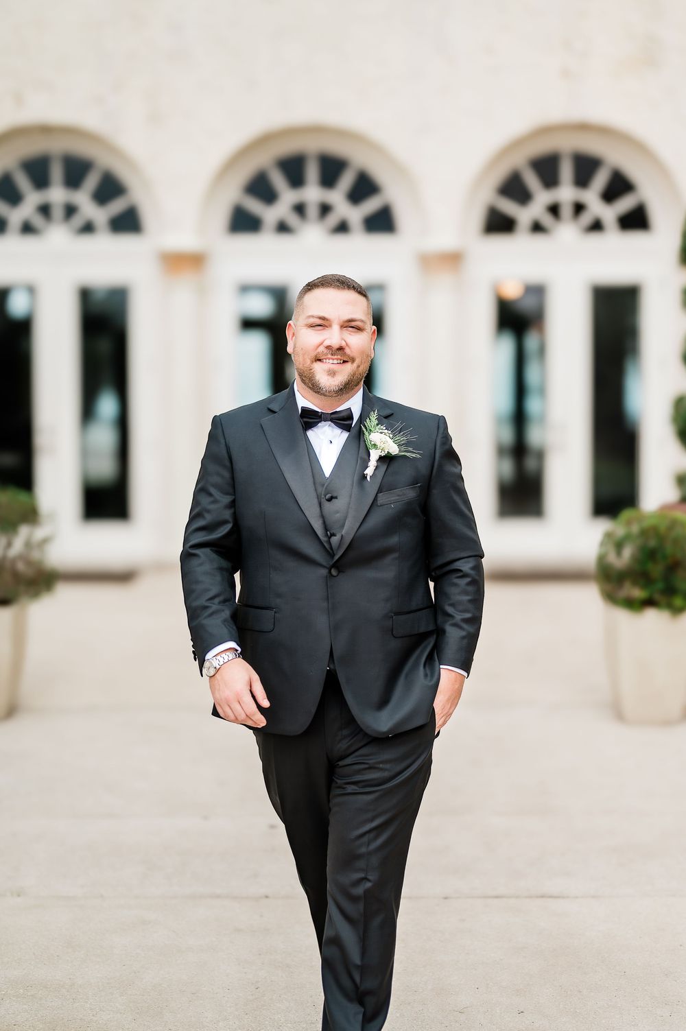A groom in a classic black tuxedo poses joyfully against a light-colored building with arched windows.