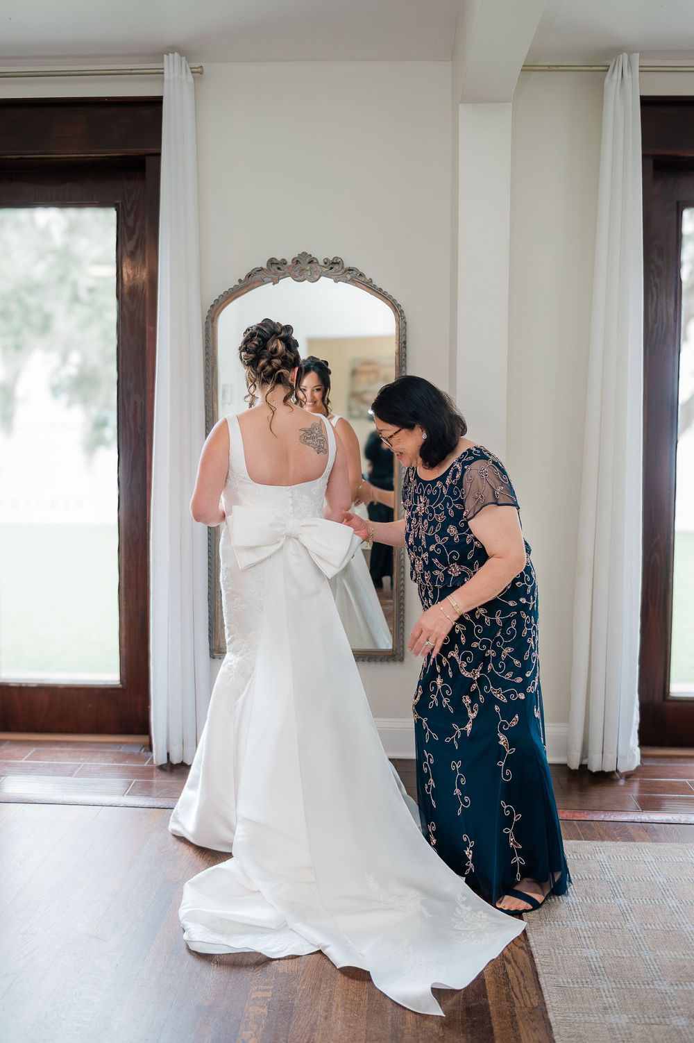 A person in a navy blue lace dress helps adjust a flowing white wedding gown in a sunlit room.