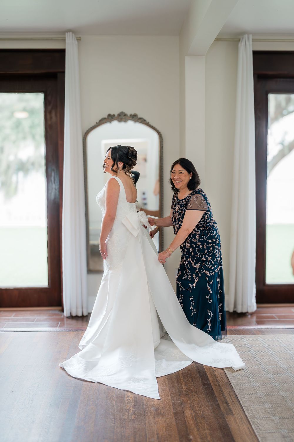 A bride in a flowing white wedding dress twirls and dances with someone wearing a navy blue dress in an elegant room.