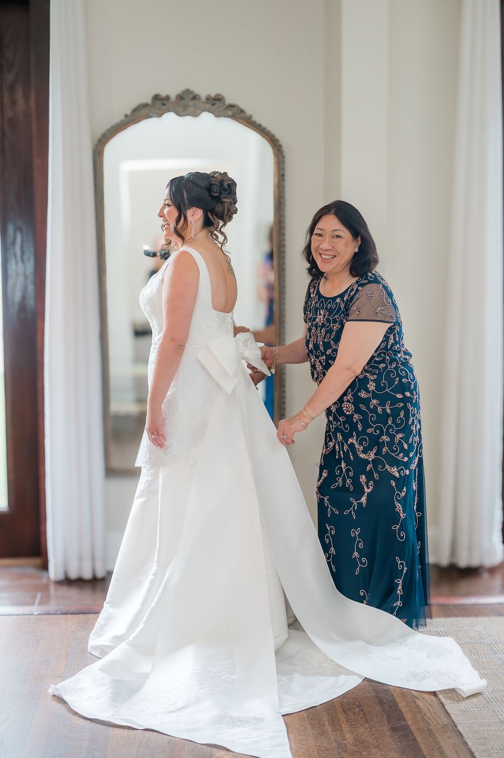 Two people in navy and white dresses help adjust a flowing white wedding gown train on hardwood floors.