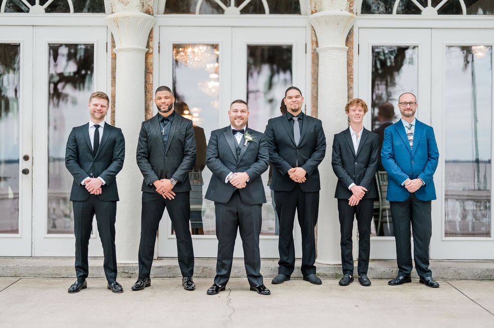 A formal group photo of groomsmen sitting on a white church bench wearing dark suits and ties.