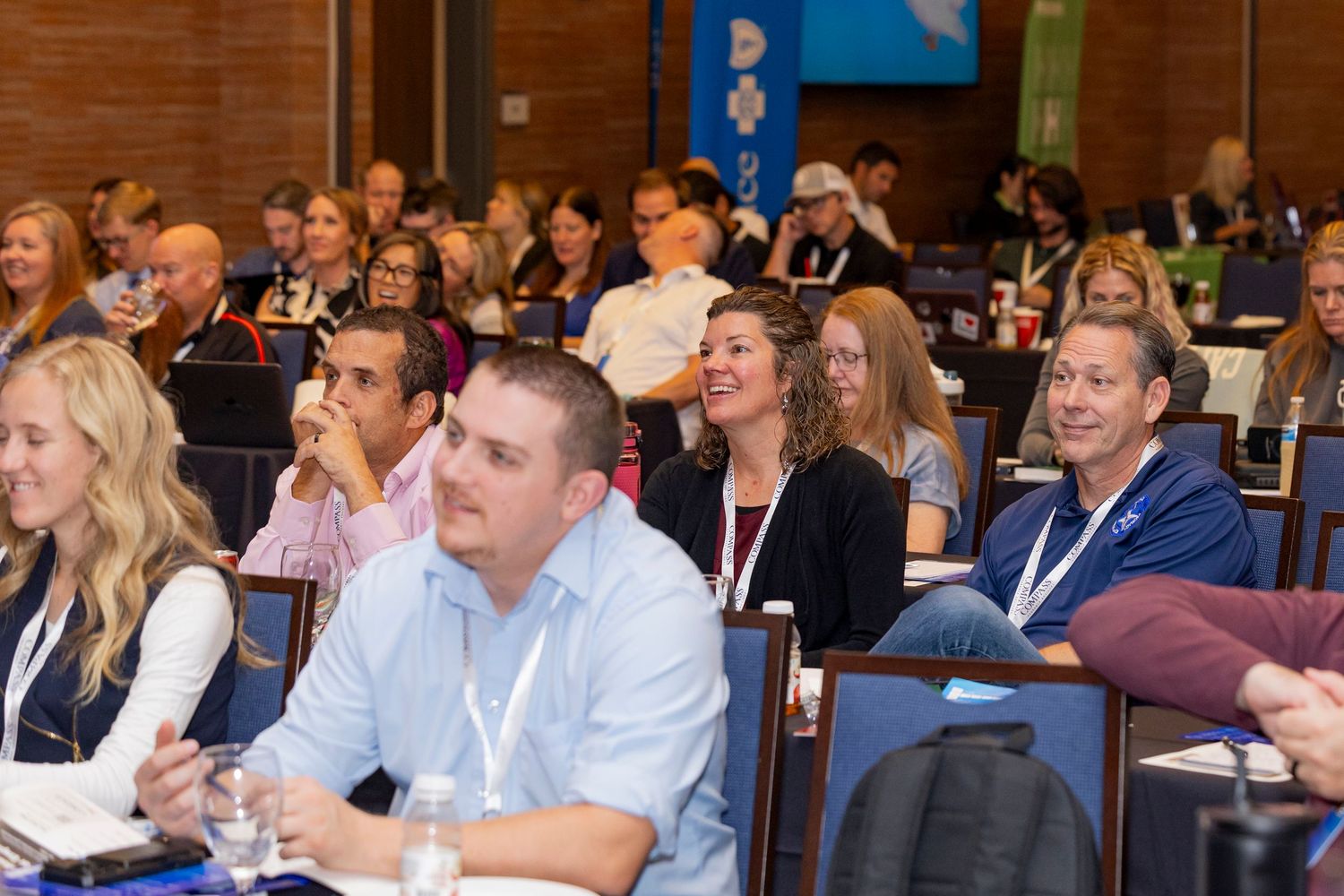 Business professionals sit at tables during a crowded conference or training session in a well-lit meeting room.