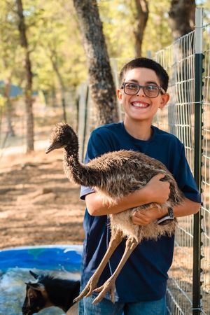 Young person in a blue shirt holds a large emu chick while smiling outdoors during his home based family photoshoot