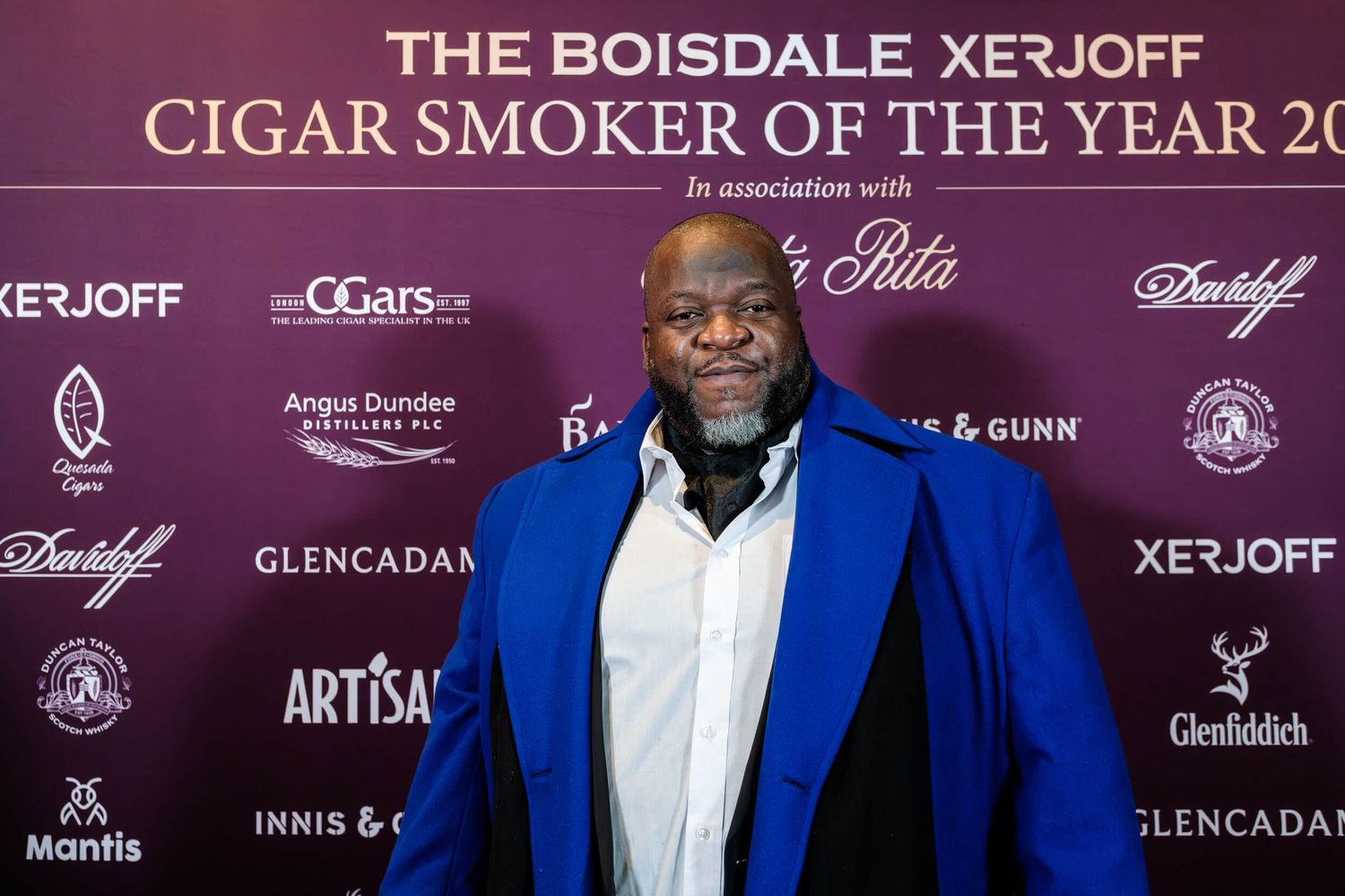 A person in a bright blue blazer and white shirt stands in front of a branded event backdrop with luxury brand logos.