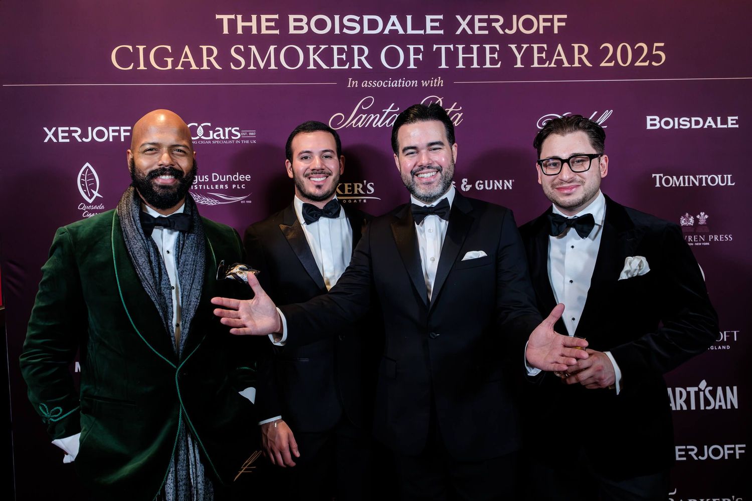 Group of people in formal black tie attire posing against a dark backdrop with event sponsor logos.