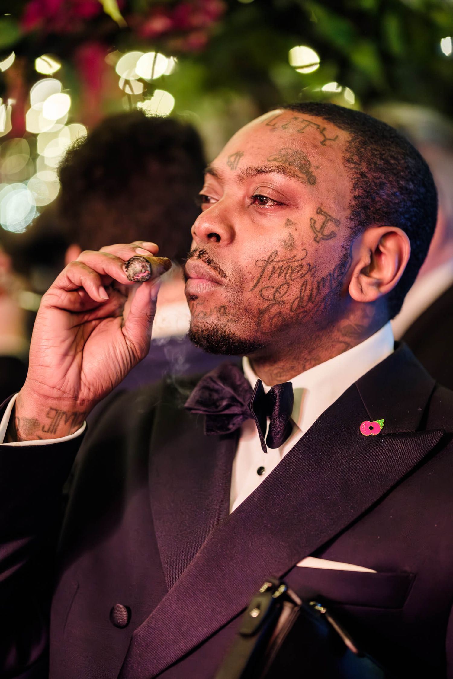 A person in a dark suit smokes a cigar while sitting in an elegant setting with bokeh lights in the background.