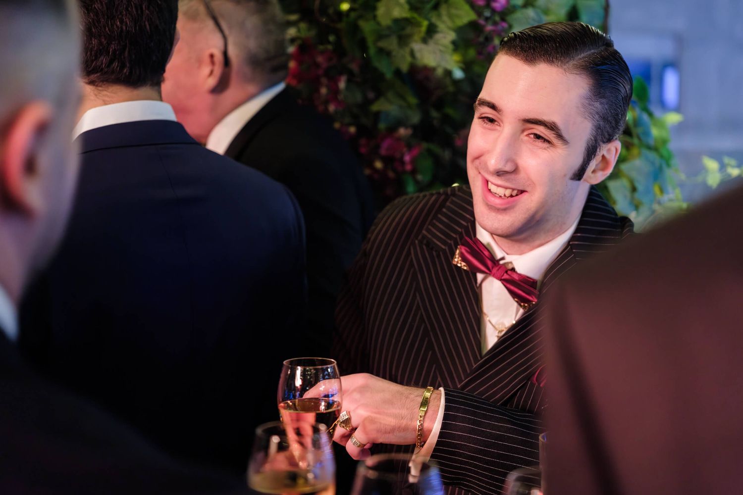 A person in a dark suit holds a glass of wine at an evening social gathering with blurred background.