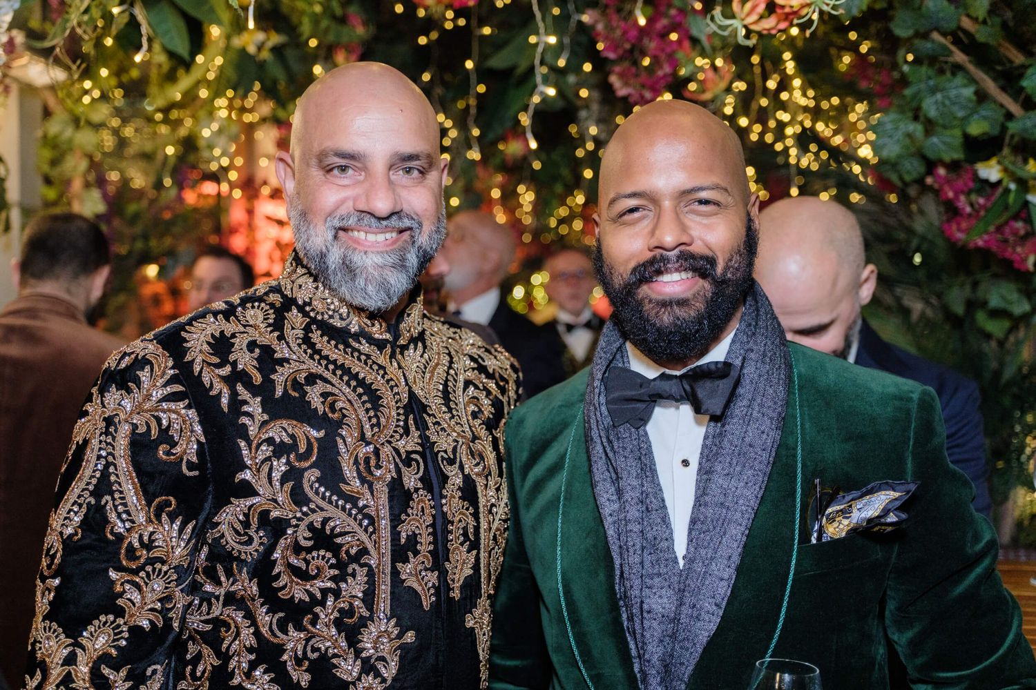 A group of people wearing formal attire including tuxedos and suits pose together at an evening event with twinkle lights.