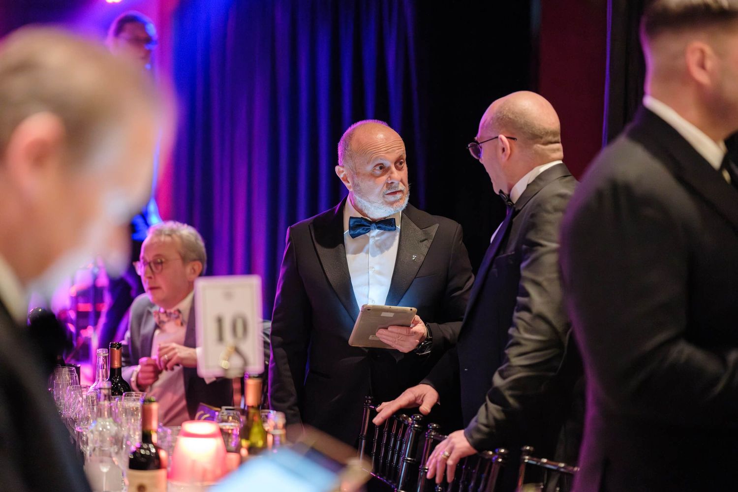 Guests in formal attire interact during an elegant evening event with purple mood lighting and table settings.