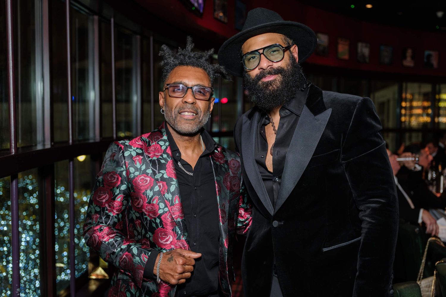 Two men pose together at an event, one wearing a floral blazer and the other in a black velvet suit.