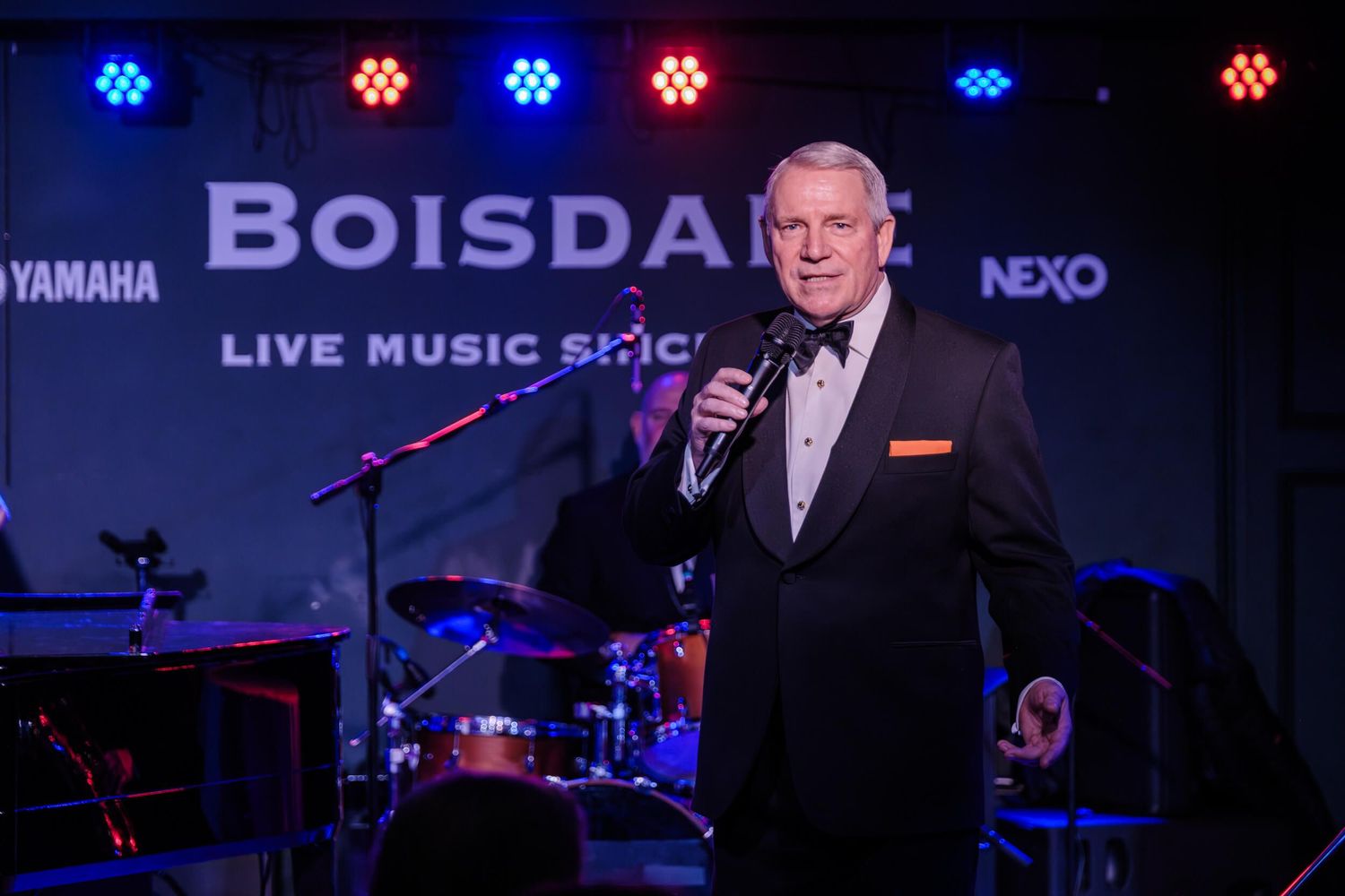 A performer in a dark suit sings into a microphone on stage with blue and red lights and Boisdale signage behind them.