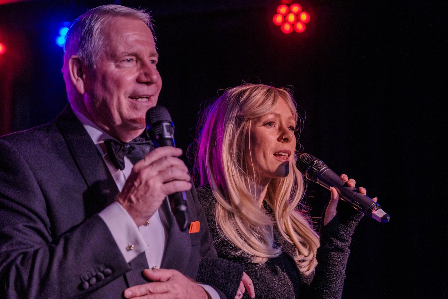 Stage performers in formal attire singing during a nighttime entertainment show with dramatic lighting.