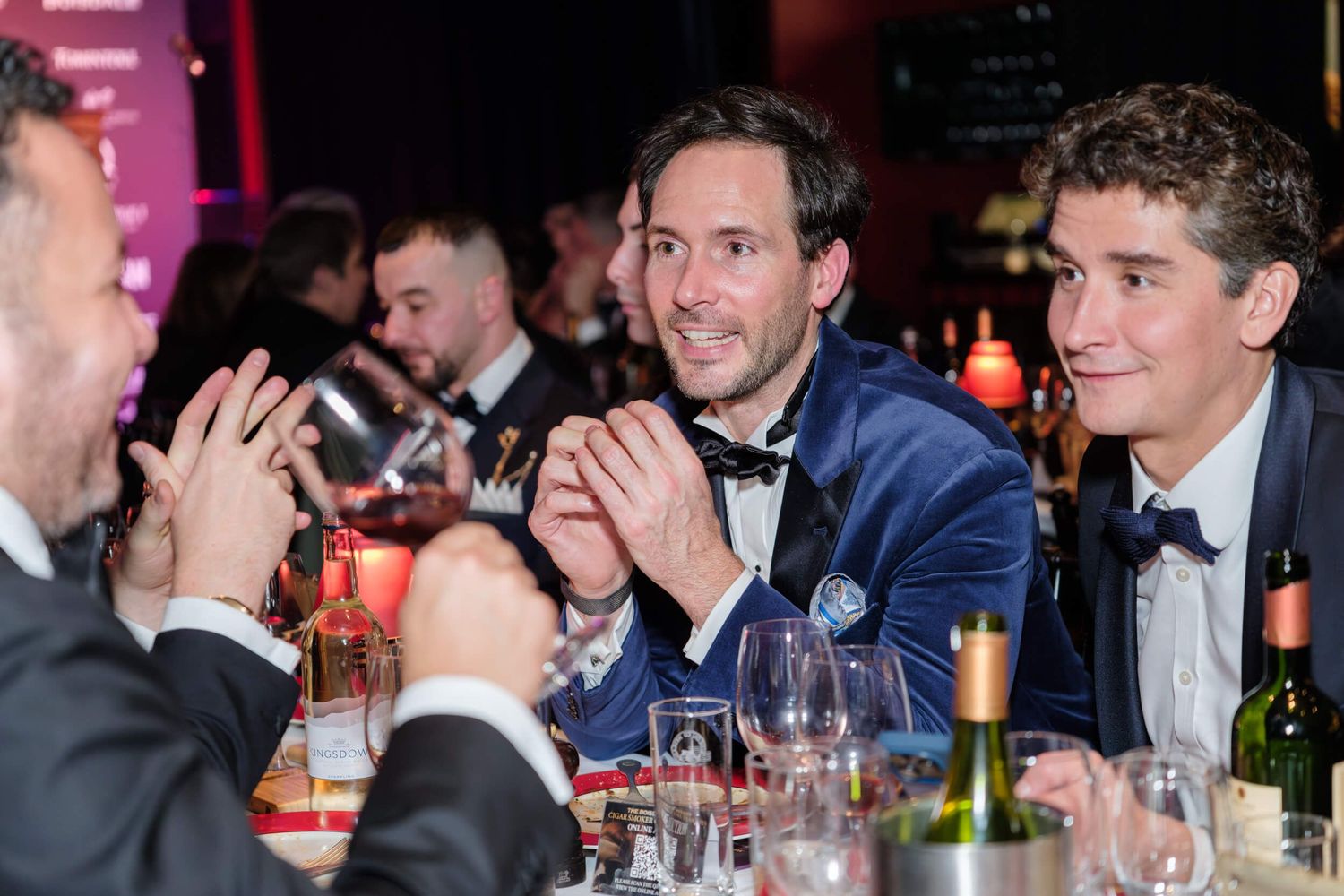 Guests seated at formal dining tables engage in lively conversation during an evening event with red lighting.