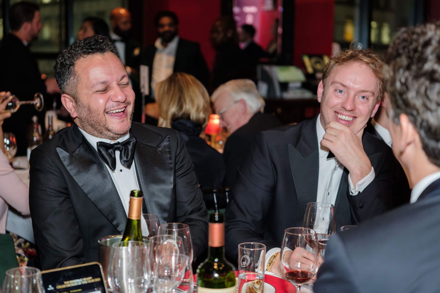 Two guests share a lighthearted moment at an elegant dinner party with wine glasses and formal attire.
