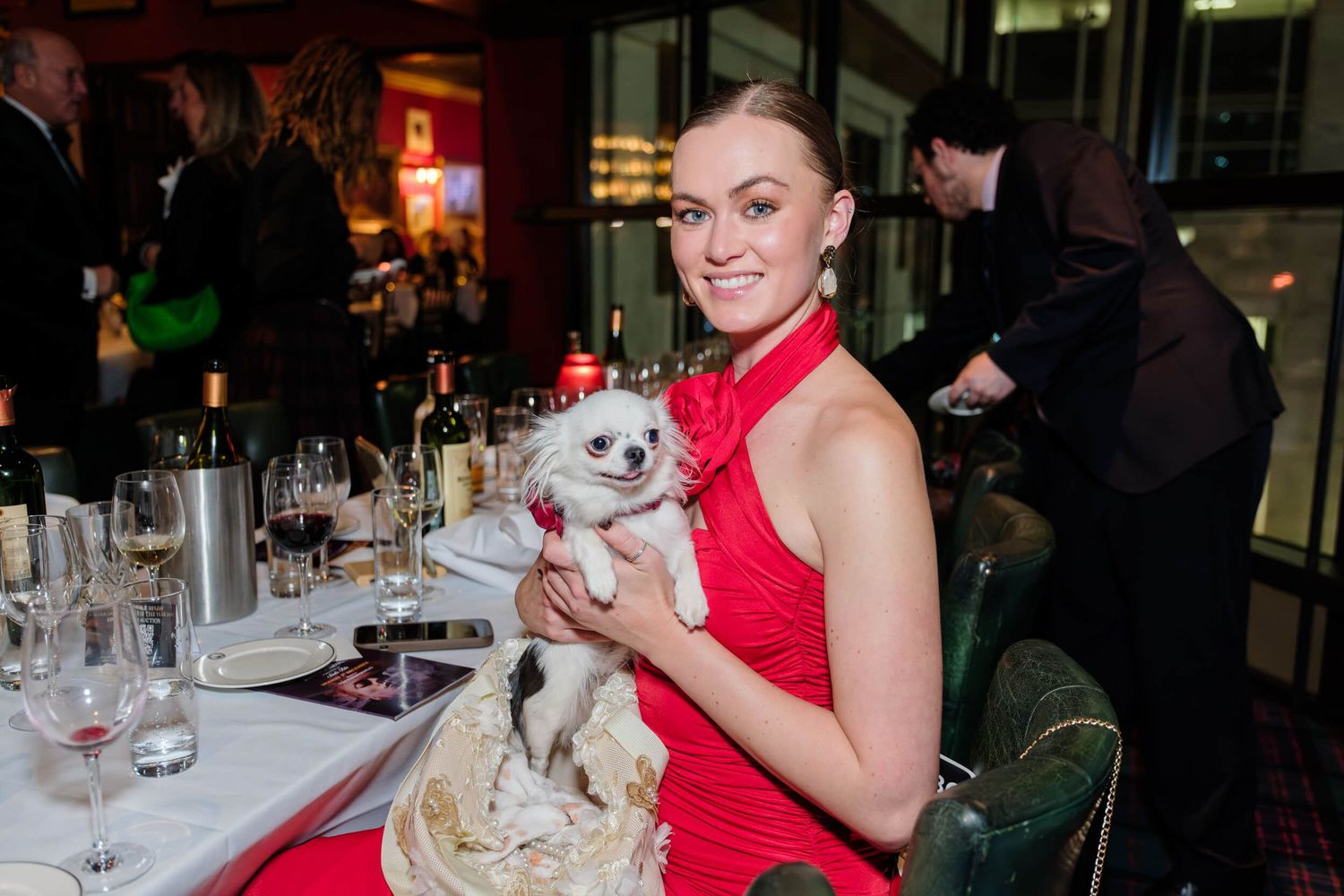 Woman in red halter dress smiles while holding small white dog at formal dinner event.