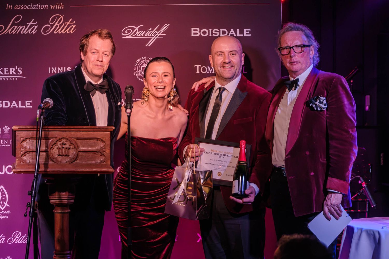 Formal event attendees gather on stage at an awards ceremony with burgundy lighting and elegant decor.