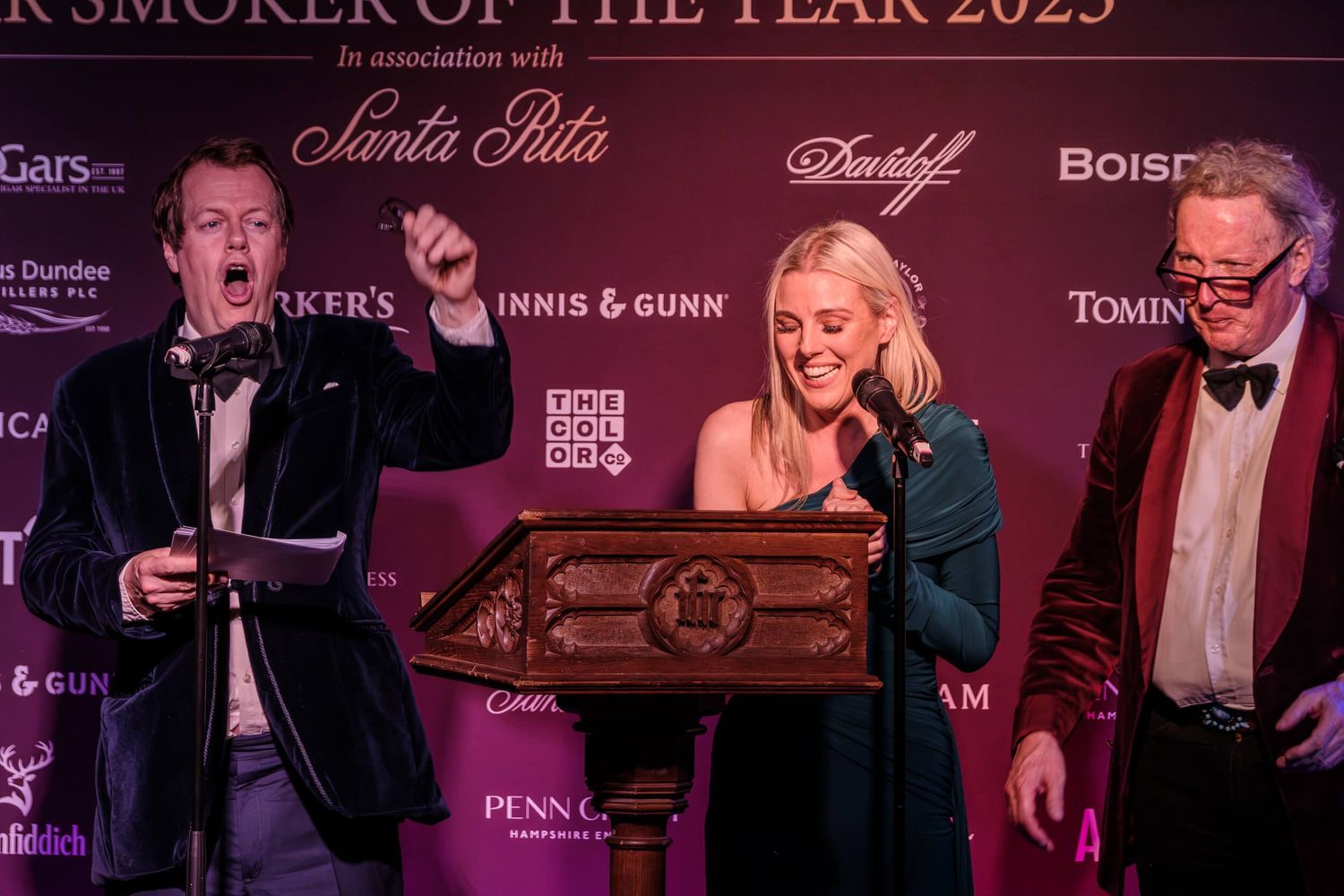 A group of people in formal attire gather at an upscale event with luxury brand logos visible in the background.