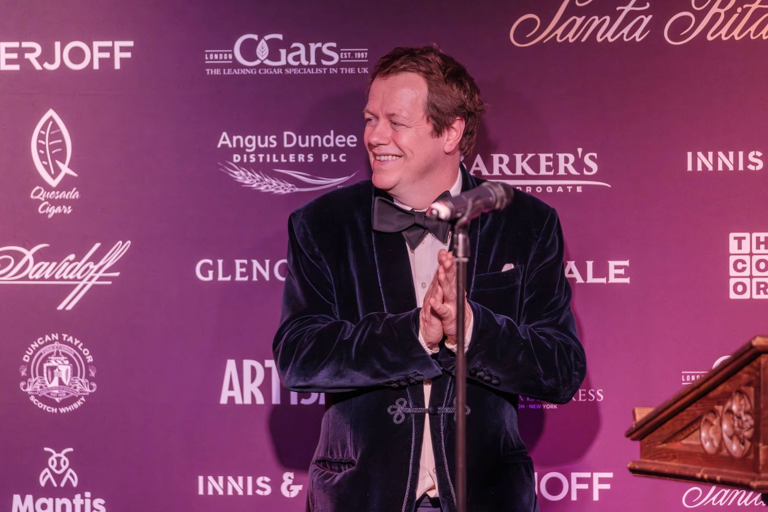 A performer in a black velvet suit jacket stands at a microphone against a burgundy backdrop with sponsor logos.