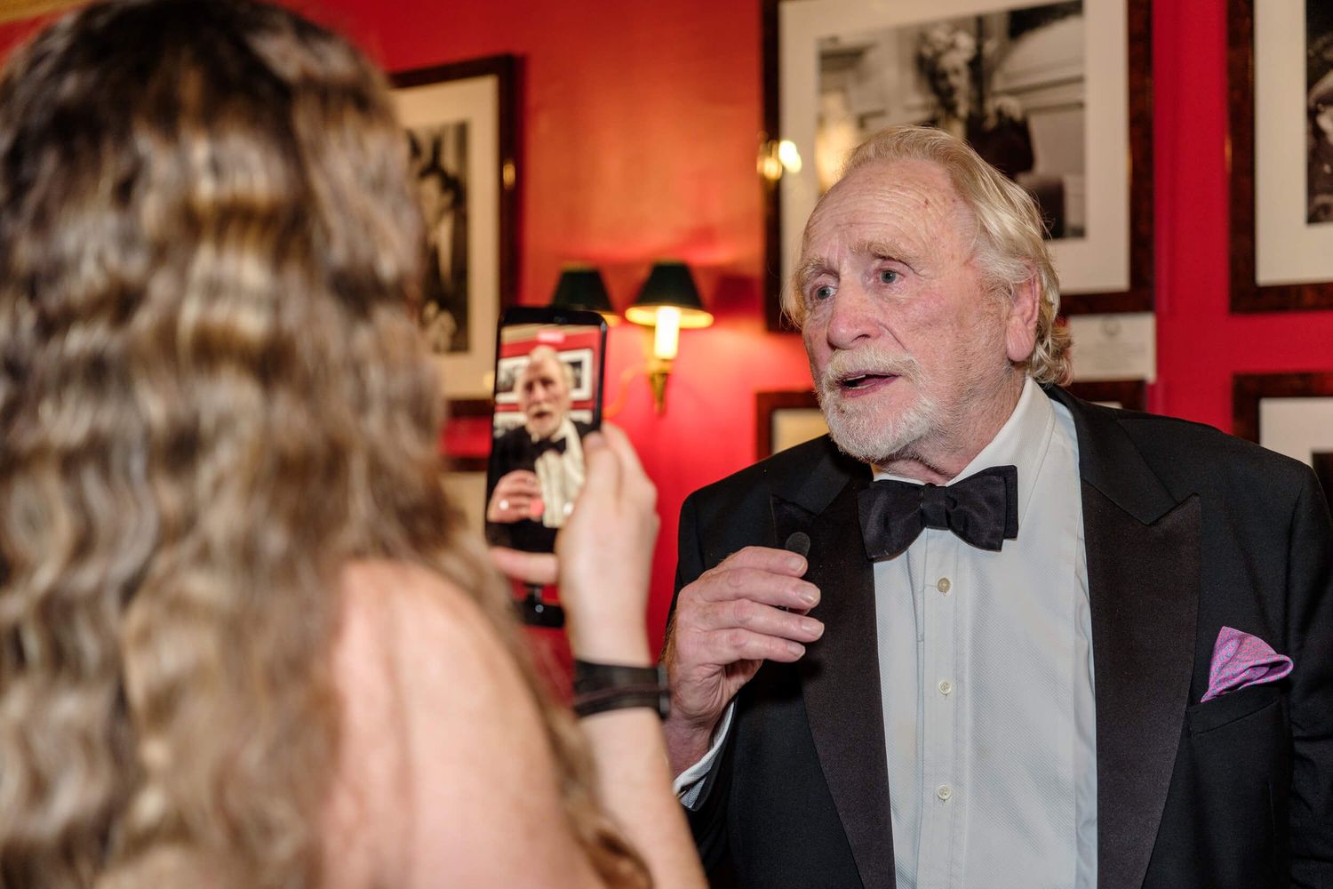 Guests in formal attire engage in conversation at an elegant evening reception with red walls and framed artwork.