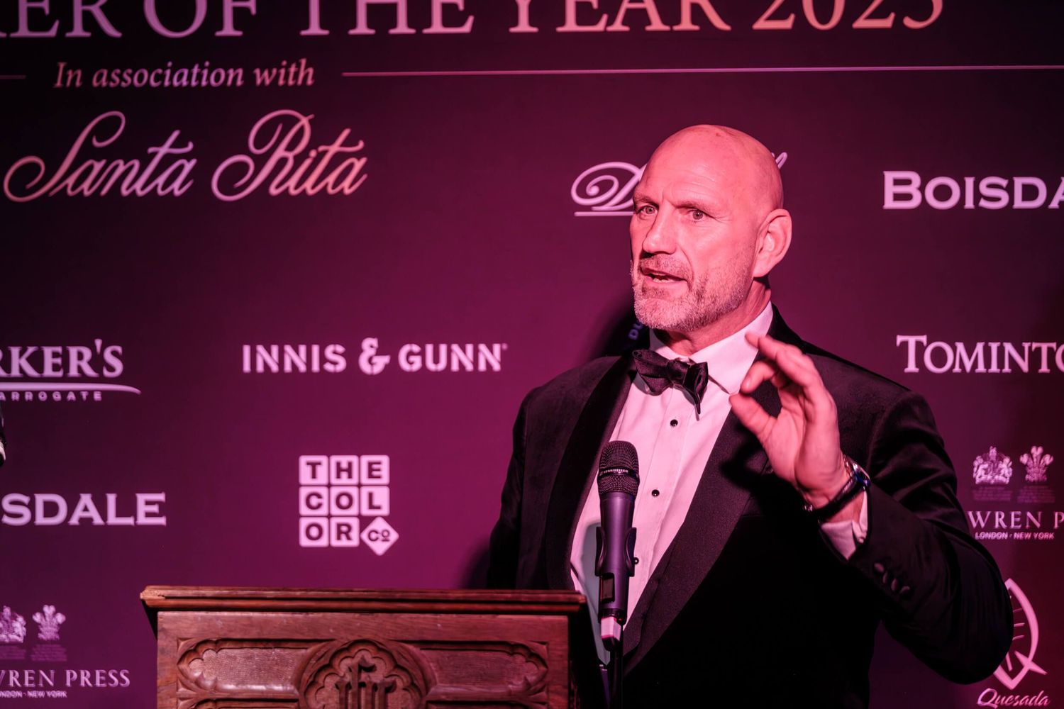 A row of wine bottles and glasses on display at an awards ceremony with purple lighting and signage.