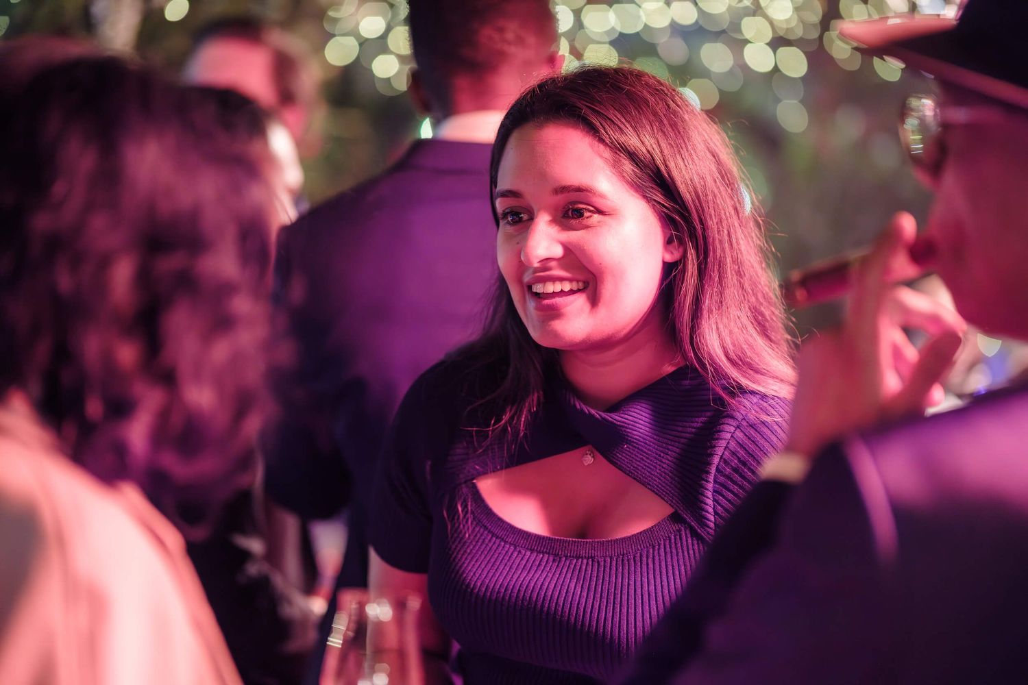 A person in a purple top smiles at a nighttime social gathering with bokeh lights in the background.