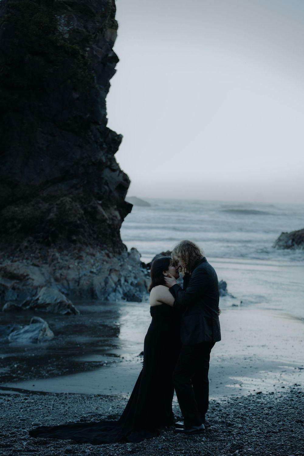 A couple shares tender moments on a rocky beach beneath towering cliffs.