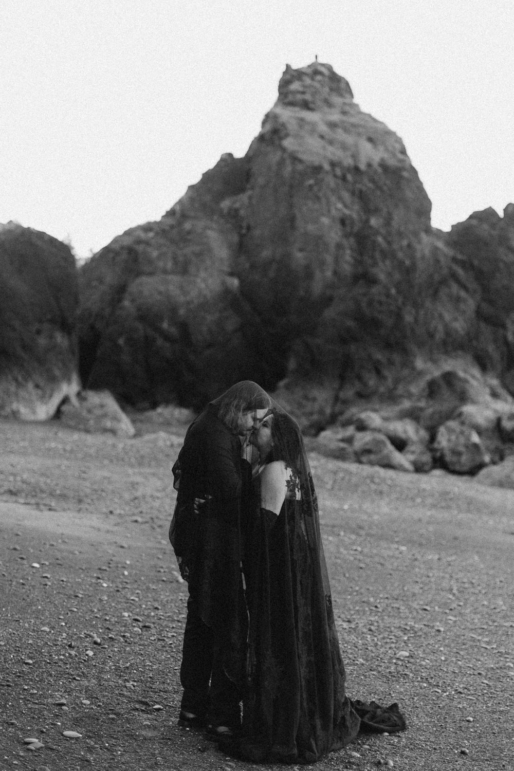 Figures in dark clothing stand against a rocky coastal landscape in dramatic black and white.