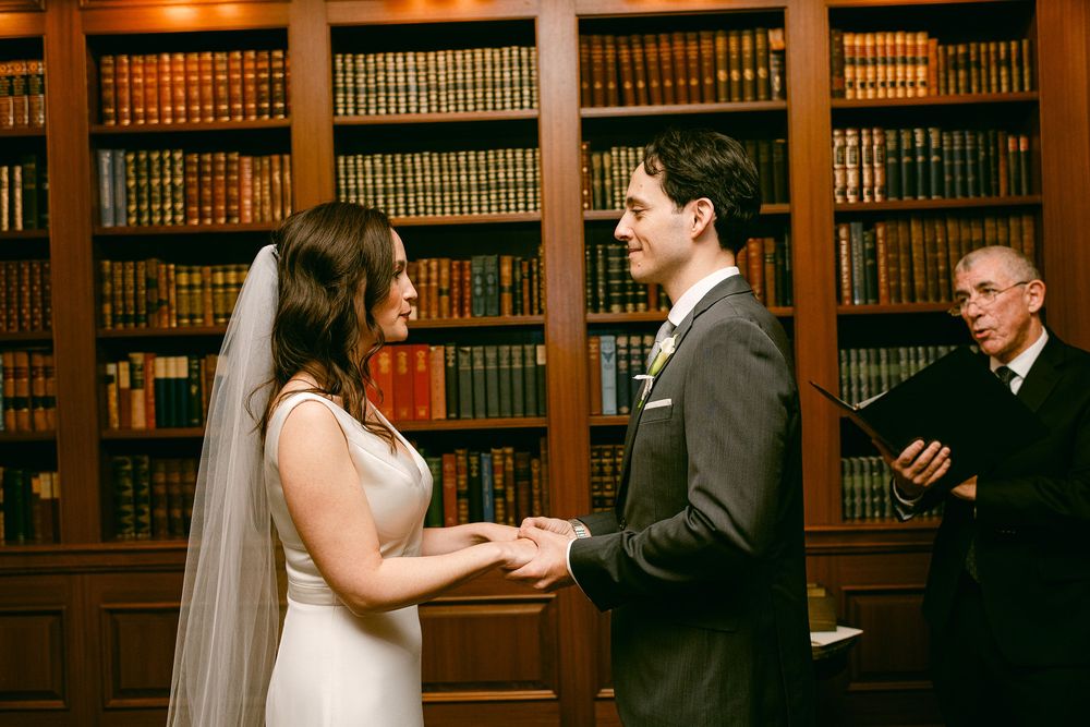 Wedding ceremony moments captured in a library setting with warm lighting and wooden bookshelves.