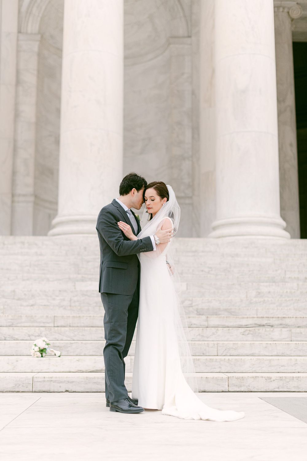 A romantic wedding couple shares an intimate moment on grand white marble steps with classical columns.