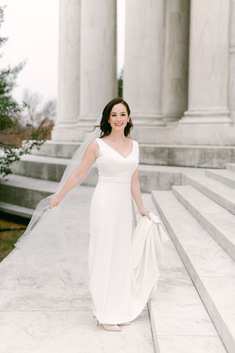 A woman in a white dress poses elegantly on marble steps with classical columns in the background.