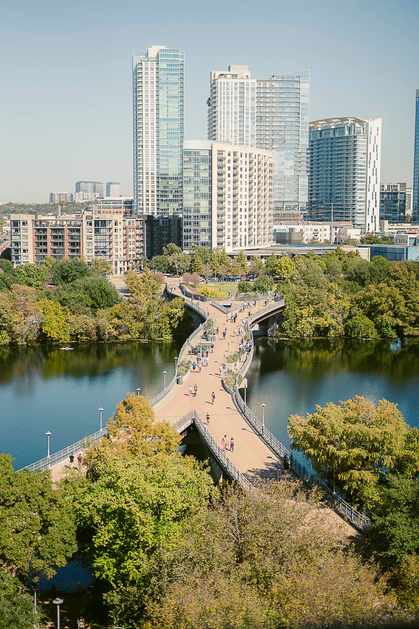 Aerial view of Lady Bird Lake pedestrian bridge with the modern Austin skyline and lush greenery along the waterfront in Austin, Texas.